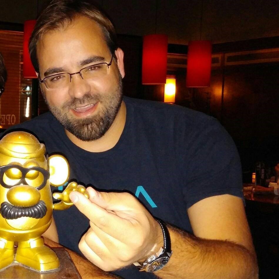 A man wearing glasses and a dark blue t-shirt smiles softly. He has dark brown hair, dark brown facial hair and is wearing glasses. The man is holding the hand of a gold-coloured “Mr. Potato Head,” which has glasses and a mustache. .