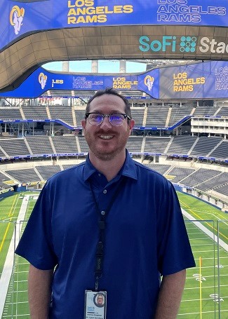 A man stands smiling into the camera. He has short, dark hair, a moustache and some stubble, and is wearing a blue golf shirt, a lanyard and glasses. Behind him there is an empty football field and empty stadium seating. The signage says “Los Angeles Rams” and “SoFi Stad.”