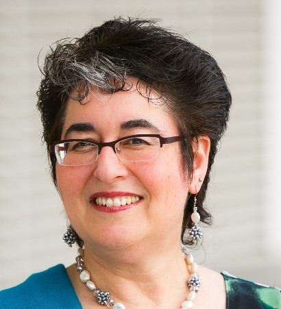 In her headshot, Betty Siegel’s head is turned slightly to the side. She is looking straight into the camera with a big smile that shows her teeth. Siegel has short, full black hair with a bit of grey at the front. She is wearing glasses, a chunky necklace, dangling earrings that complement her necklace, and a green, blue and black shirt.