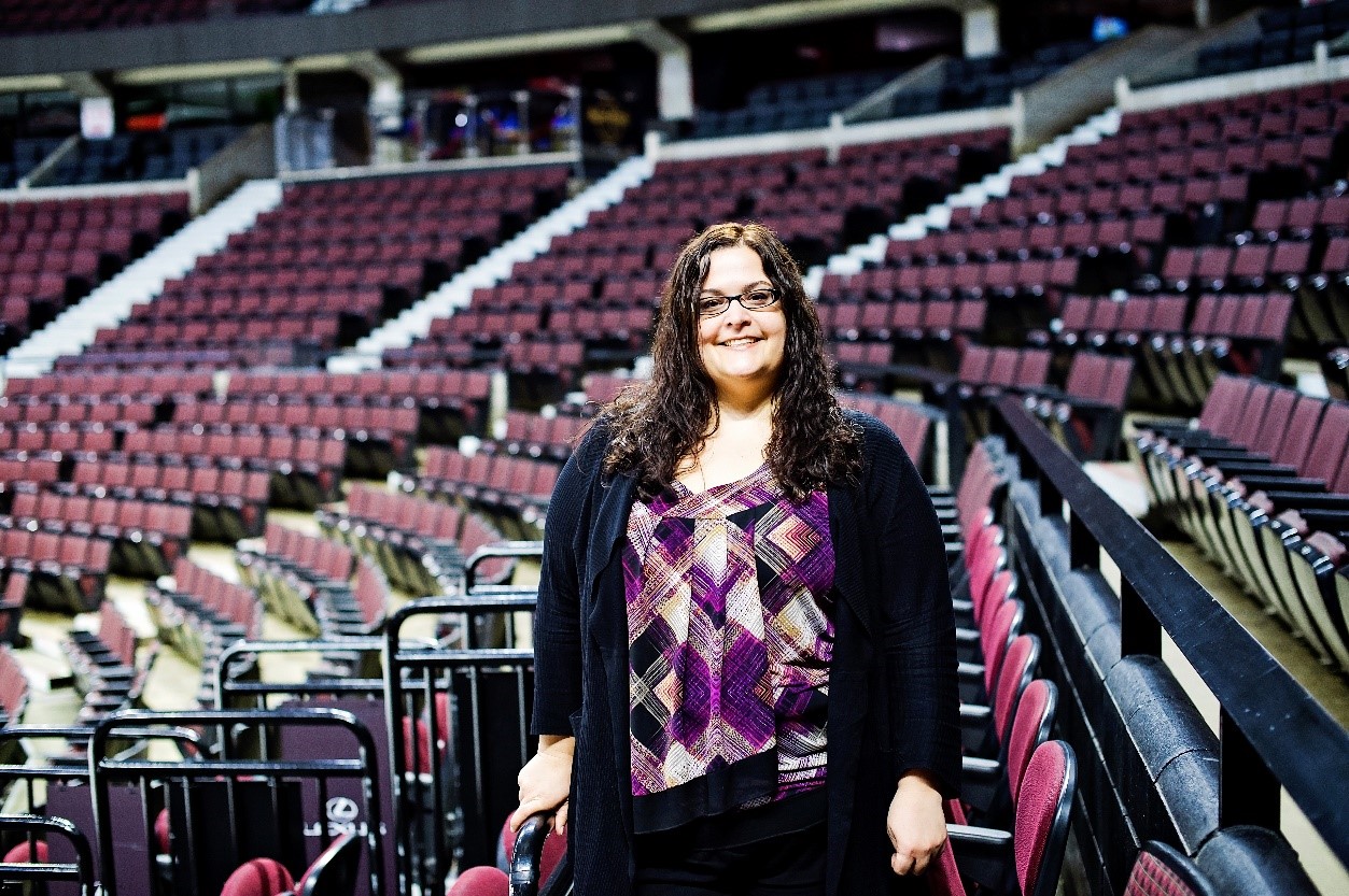 A woman stands smiling in front of rows of empty seats in an arena. She has long, dark curly hair and is wearing glasses, a colorful patterned top, black sweater and black pants.