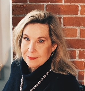In her headshot, Deirdre’s head is slightly tilted and she has a soft smile. Her hair is blond and flows past her shoulders. She is wearing a black cowl-neck sweater, chunky necklace and earrings. There is a red brick wall in the background.