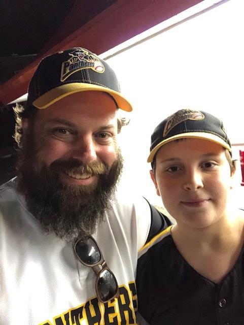 A man with dark hair, a moustache and beard stands smiling wearing a white jersey that says “Panthers” and a black and yellow Panthers cap. Beside him, a boy is wearing the same baseball cap and a black jersey.