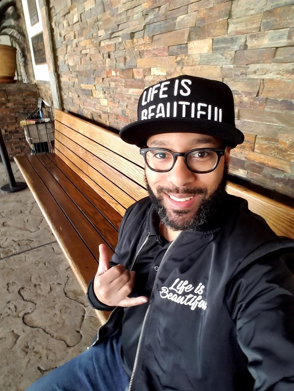 Geo is sitting on a long wooden bench in front of a brick wall. He has light brown skin, a moustache and beard. His black hat and jacket both say “Life is Beautiful.”