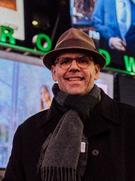 A smiling man wearing glasses and a hat stands in front of large projection screens.